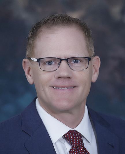 Jard Brown; A person in a suit and tie smiles confidently at the camera. They are wearing glasses and have short hair. The background is a soft, blurred mix of dark colors, adding to the professional tone.