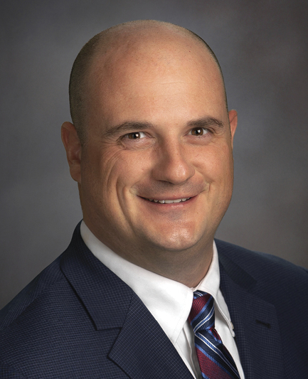 Darren Gragg; A bald man in a dark blue suit and red striped tie smiles confidently against a neutral gray background. Professional portrait with a warm tone.