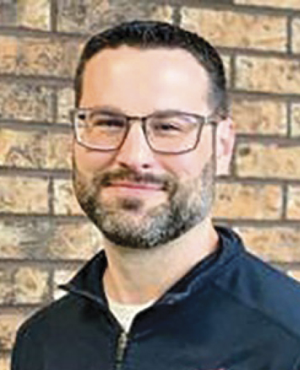 Taylor Hight; Man with dark hair and beard, wearing glasses and a dark collared shirt, smiles softly in front of a textured brick wall.