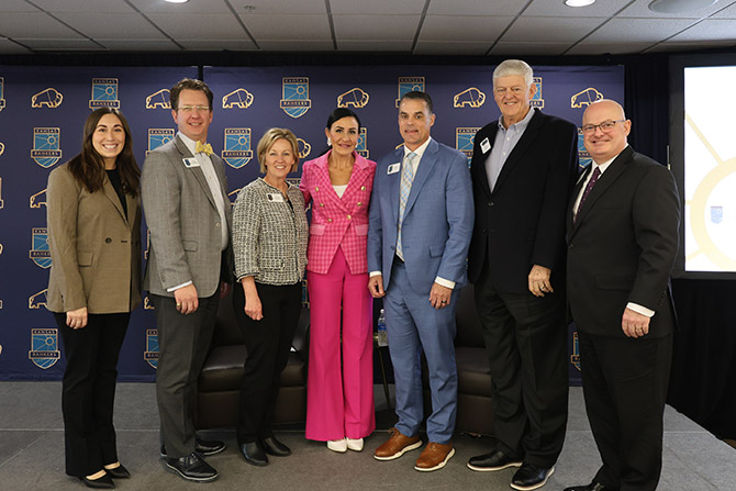 A group of seven people, both men and women, are standing together, smiling. They are dressed in formal attire. Behind them is a backdrop with a blue and orange logo featuring a bear, suggesting a team or organization setting. The atmosphere is professional and friendly.