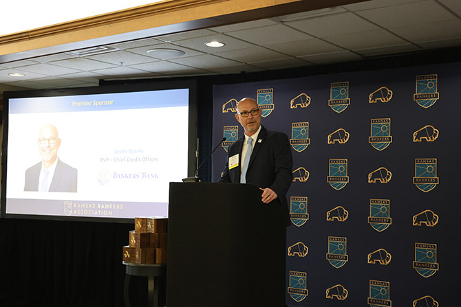 A man in a suit speaks at a podium against a backdrop with Kansas Bankers Association logos. A screen displays a business presentation.