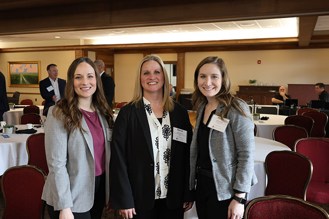 Three women dressed in business attire stand smiling in a conference room with round tables and attendees in the background. They appear professional and friendly.