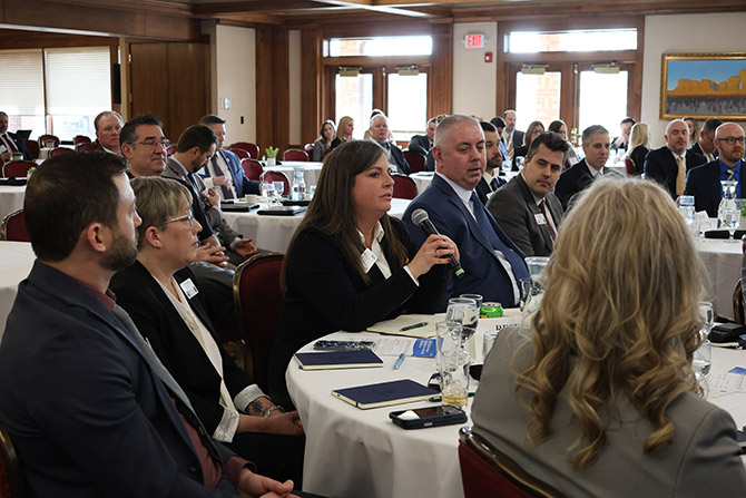 A woman speaks into a microphone in a conference room filled with people in business attire, seated at round tables. The mood is focused and engaged.