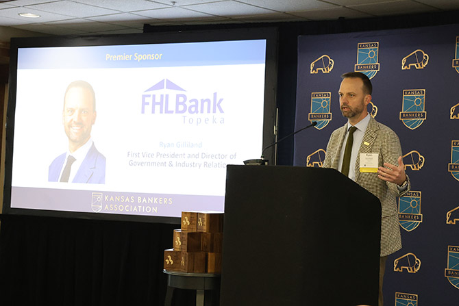 A man stands at a podium speaking, next to a large screen displaying a presentation slide for FHL Bank Topeka, a premier sponsor, during a Kansas Bankers Association event.