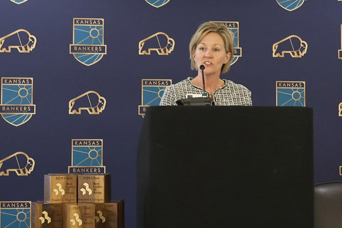 A woman speaks at a podium with a Kansas Bankers logo backdrop. Multiple plaques are stacked beside her, suggesting an awards event.