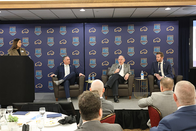 Four individuals in business attire participate in a panel discussion on stage. Behind them is a blue backdrop with logos, and the audience is in the foreground.