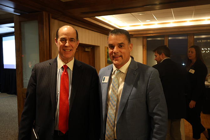 Two men in suits stand side by side at an indoor event. One wears a red tie, the other a light blue tie, both smiling. A few people converse in the background.