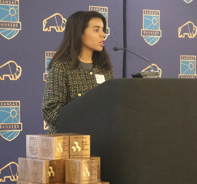 A woman speaks at a podium with a microphone, set against a backdrop with "Kansas Bankers" logos. Wooden award blocks are stacked nearby. Professional tone.