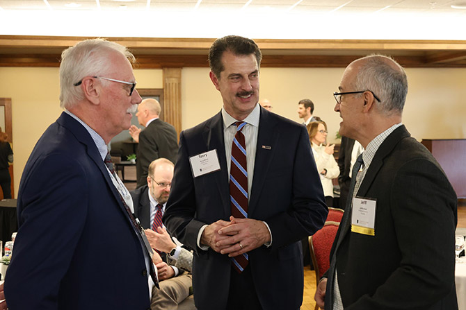 Three men engage in conversation at a formal event, wearing suits and name tags. The atmosphere is professional, with others mingling in the background.