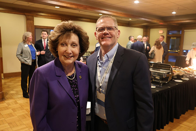 A woman in a purple blazer and a man in a suit, both smiling, stand at an indoor event with people socializing and buffet tables in the background.