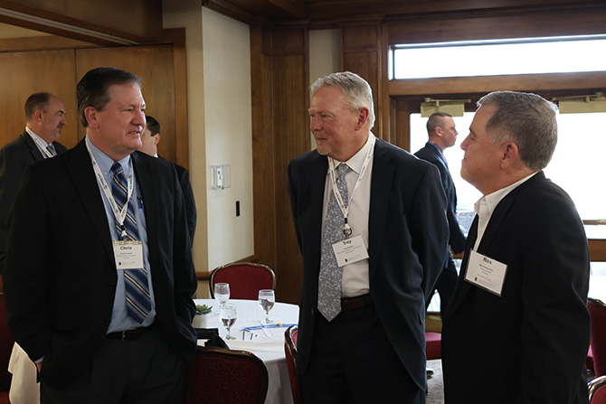 Three men in suits engaged in conversation at a formal event. They wear name tags and stand in a room with tables, red chairs, and wooden paneling.