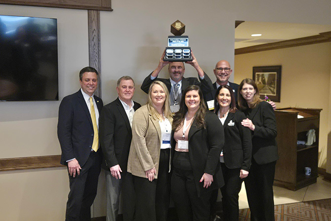 A group of people in professional attire smile together while one man in the back holds up a trophy.