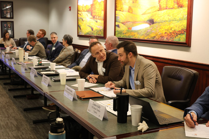 A group of people sit around a conference table in discussion. Papers and drinks are on the table. A landscape painting hangs on the wall.