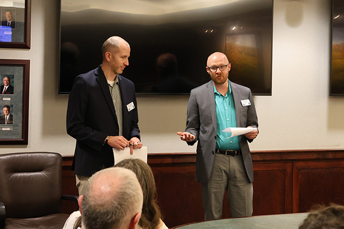 Two men stand in a conference room, speaking to an audience. They hold papers and are dressed in business attire. The mood is professional and engaged.
