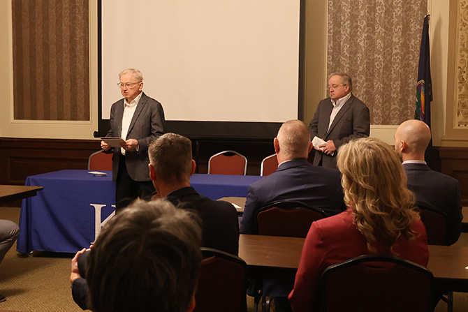 Two men stand at the front of a conference room with a blue tablecloth. One speaks to an audience seated at tables, creating a formal and attentive atmosphere.