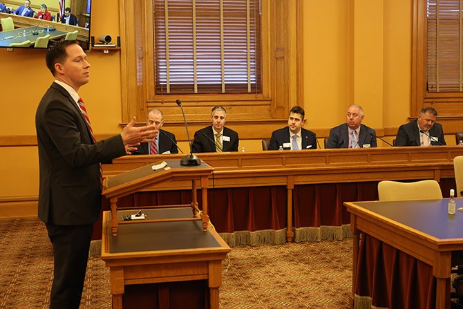 A man in a suit speaks at a podium in a wood-paneled room. Five men in suits sit attentively at a long table. A screen displays the room's view.