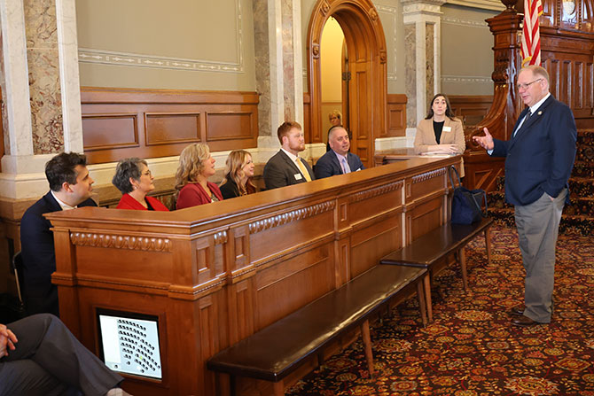 A man stands and speaks to a seated group of people in a wood-paneled room with ornate decor, marble columns, and an American flag.