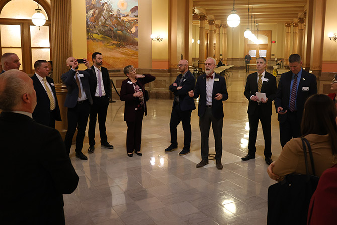 A group of professionals in formal attire stand in a grand hallway with ornate decor. One man speaks animatedly, suggesting a discussion or tour.