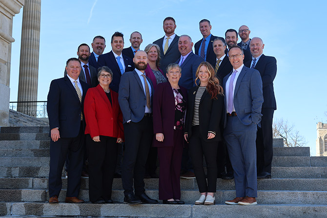 A group of seventeen professionally dressed adults stands on outdoor steps in front of a building, posing for a group photo on a sunny day.