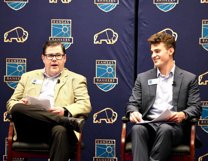 Two men in suits sit on stage with Kansas Bankers backdrop. One speaks, the other listens and smiles. Professional and engaging atmosphere.