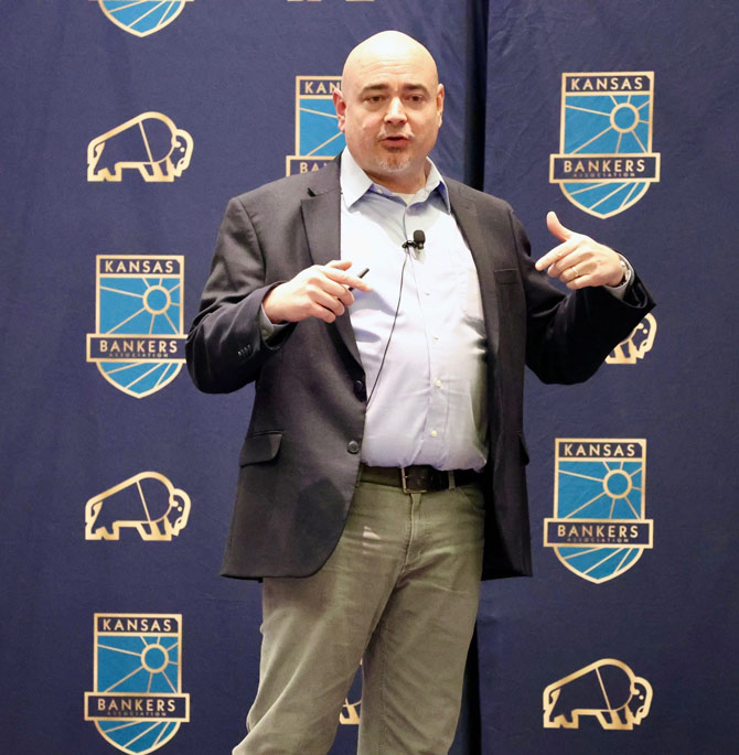 A man in a suit gestures while speaking on stage, in front of a Kansas Bankers backdrop with buffalo logos, conveying a professional tone.