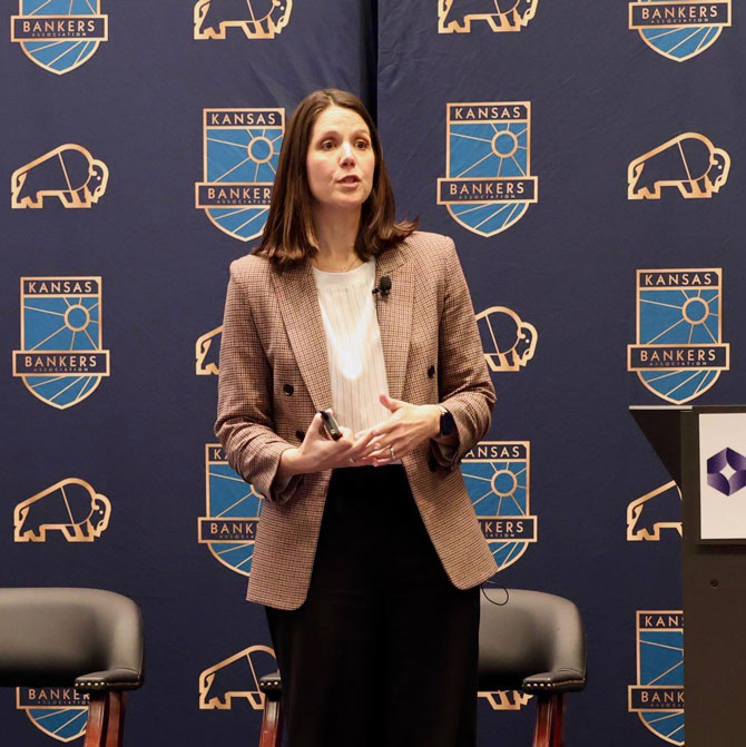 A woman in a plaid blazer speaks confidently on stage, holding a remote. Behind her is a "Kansas Bankers" logo on a blue backdrop.