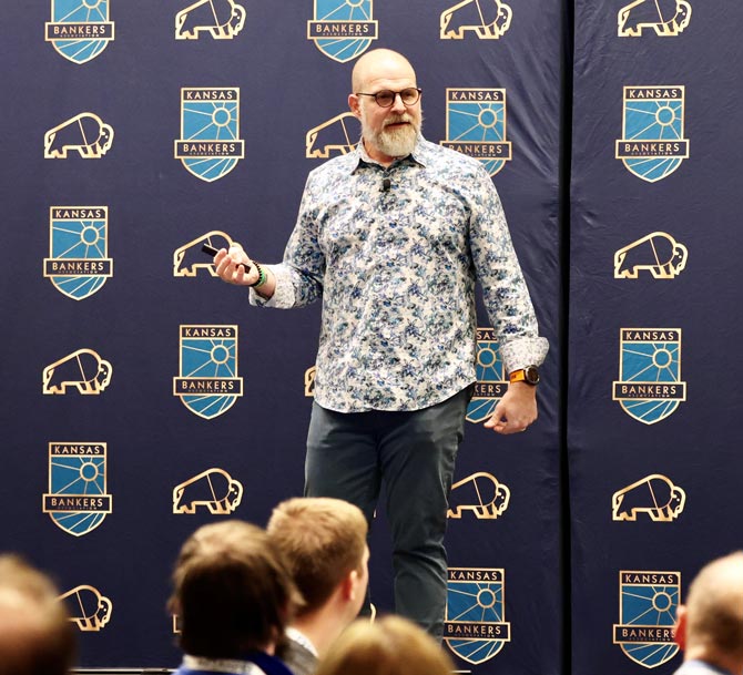 A speaker in a floral shirt presents energetically at a Kansas Bankers event. The backdrop features the event logo and icons of bison.