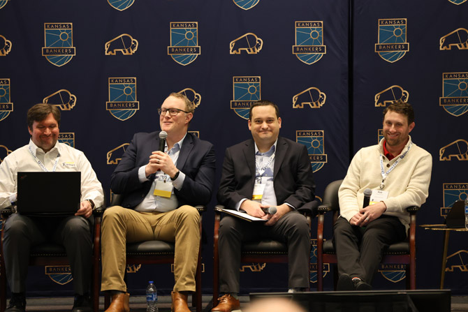 Four men sit on a panel stage with a "Kansas Bankers" backdrop. One holds a microphone, engaged in discussion, creating a professional atmosphere.