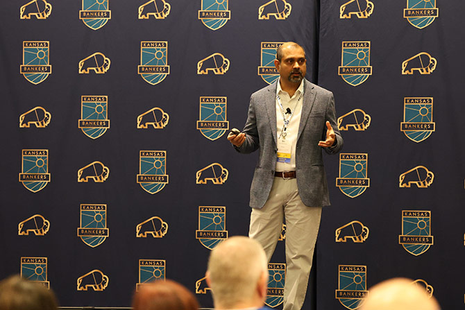 A speaker in a suit gestures on stage at a conference. The backdrop features Kansas Bankers logos and bison icons, creating a formal tone.