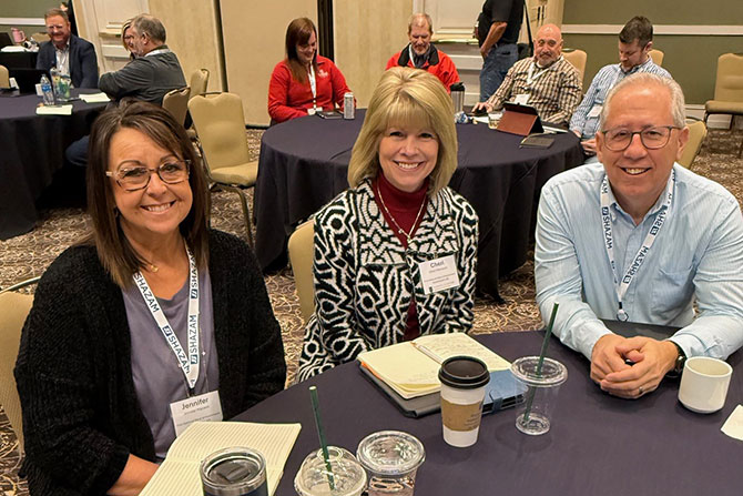 Two women and one man smile while sitting at a table with badges on with other people at tables behind them.