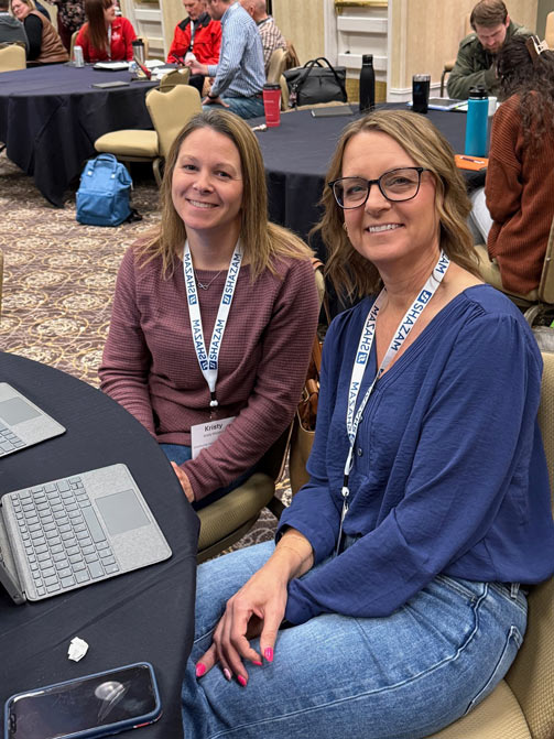 Two women smiling at a conference, seated at a table with laptops. They wear name badges and casual attire. A busy, social atmosphere surrounds them.