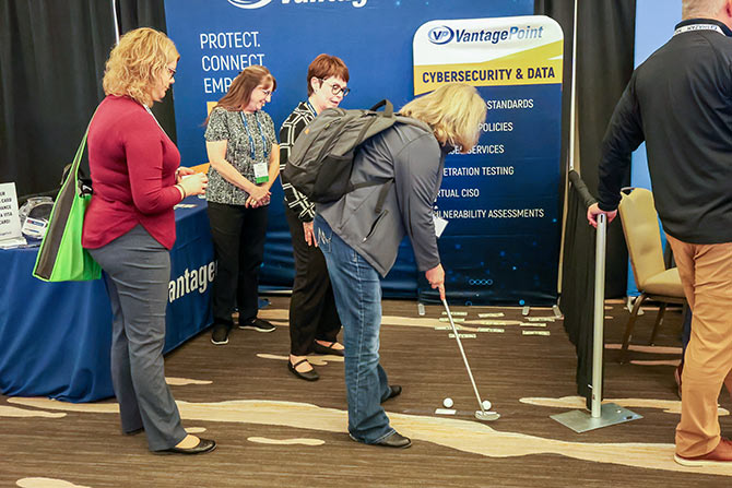 People at an event booth engage in mini-golf. A woman putts, while others watch. The backdrop features cybersecurity and data signage.