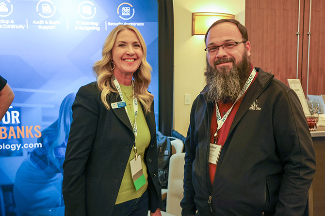 Two people smile at a conference, standing in front of a blue booth with text about technology and security. Both wear conference lanyards, conveying a professional and friendly atmosphere.