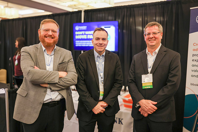 Three men in business attire stand smiling at a conference booth. The backdrop features a sign and displays, suggesting a professional event setting.