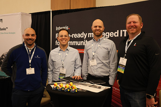 Four men smiling, standing at a trade show booth for a managed IT service. The booth features branding and promotional materials on a table.
