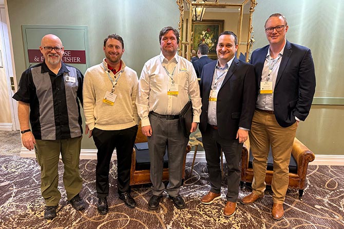 Five men stand in a warmly lit room, smiling, wearing business casual clothing and conference badges. A mirror and painting are in the background.
