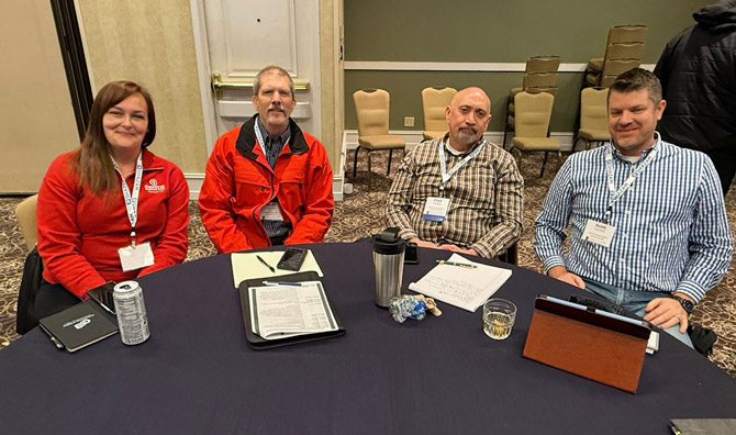 One woman and three men smiling at a conference, seated at a table with notes and electronics. They wear name badges and professional attire.