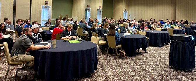 A large group of people sits attentively at round tables in a conference room, listening to a presentation. The atmosphere is focused and engaged.