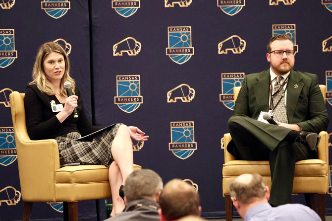 A woman and a man sit on stage, engaged in discussion at a Kansas Bankers event. She holds a microphone, and both wear formal attire with a backdrop of logos.
