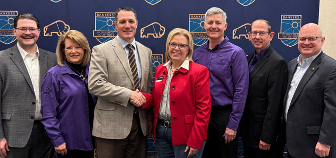 A group of seven people standing in front of a "Bankers Healthcare Solutions" backdrop. Two people in the center shake hands, smiling.