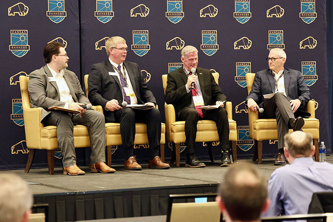 Four men sit on stage chairs, engaged in a panel discussion against a backdrop with the Kansas Bankers Association logo, conveying a professional tone.