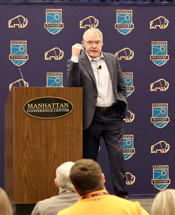 Man speaking energetically at a podium labeled "Manhattan Conference Center," with Kansas Bankers logos on backdrop, addressing an audience.