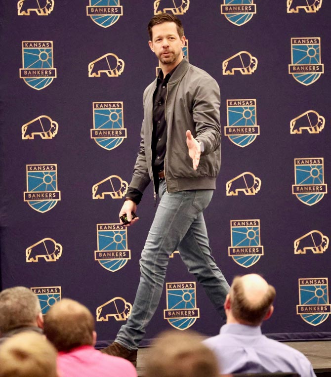 Man on stage in a casual jacket and jeans speaks to an audience. The backdrop features "Kansas Bankers" logos, conveying a professional tone.