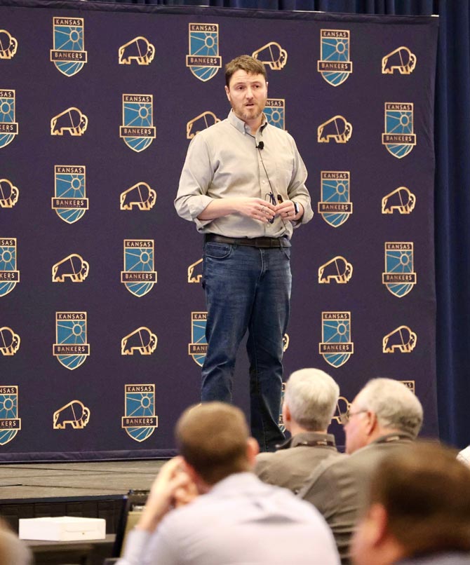 Man on stage in a button up shirt and jeans speaks to an audience. The backdrop features "Kansas Bankers" logos, conveying a professional tone.