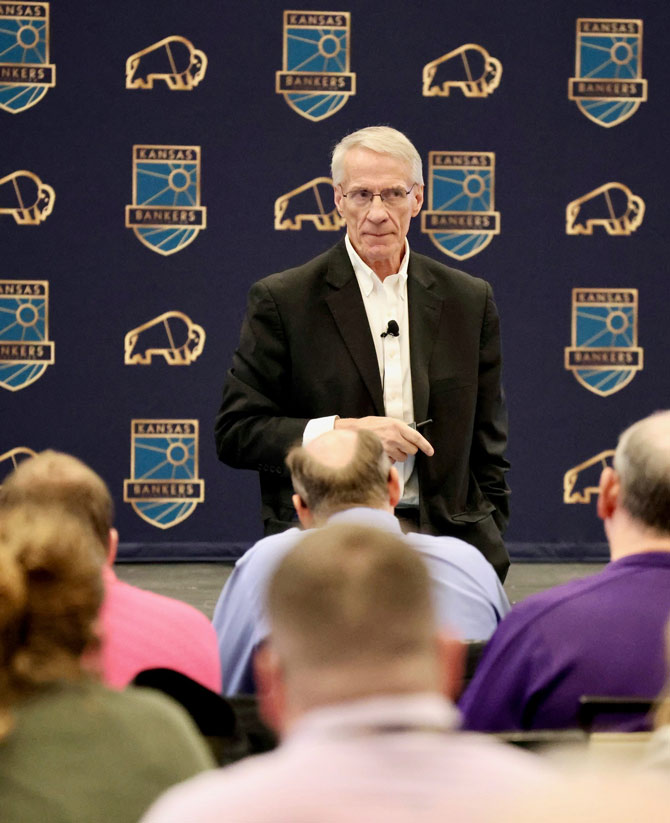 Man on stage in a black suit looks over the audience. The backdrop features "Kansas Bankers" logos, conveying a professional tone.