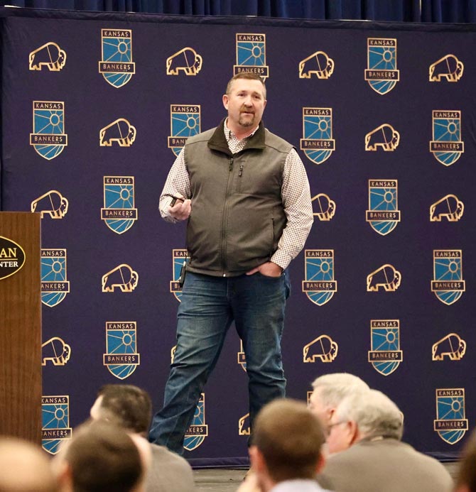 Man on stage in a button up shirt, vest and jeans speaks to an audience. The backdrop features "Kansas Bankers" logos, conveying a professional tone.