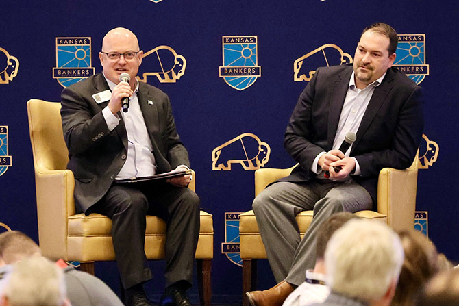 Two men in suits speak into microphones while seated on stage at a Kansas Bankers event. A backdrop displays the event logo, suggesting a formal discussion.