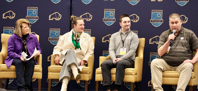 A panel of four people seated on stage at a Kansas Bankers event. One person speaks into a microphone. The backdrop displays logos.