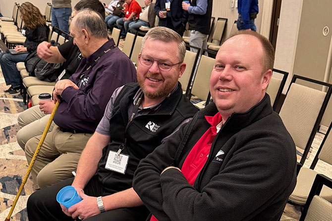 Two men smiling at a conference, seated among others. They wear casual business attire and look engaged. The atmosphere is friendly and relaxed.