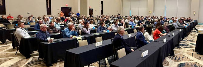 A large conference room filled with attentive attendees seated in rows facing the front. Tables are covered in black cloths, and name cards are visible.